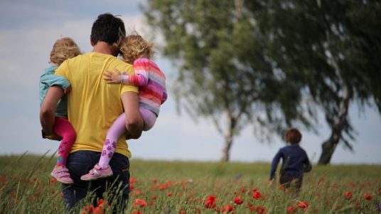 A man holding two kids in a field with red flowers next to a big tree