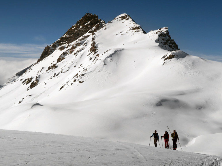 three people walking on the snow at the foot of a rocky mountain