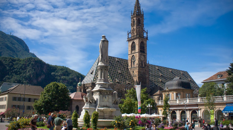 the church of bolzano with its green and gold roof and the fountain with flowers in the middle of the square